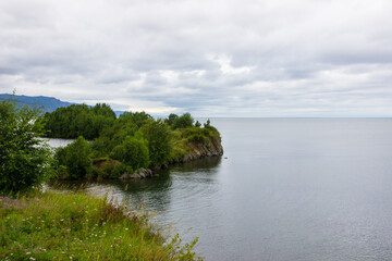 Fototapeta na wymiar Small rocky islands covered with green grass and green trees on the left. Grey rippling water on the right. A path of dark clouds in a light cloudy sky