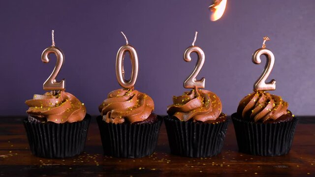 Happy New Year's Eve 2022 chocolate cupcakes decorated with gold burning candles on aqainst dark wood table setting background. Lighting candles.