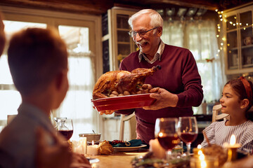 Happy senior man serves Thanksgiving turkey to his family at dining table.