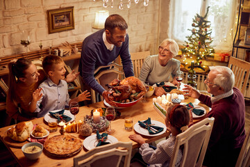 Happy man serves roast turkey for his family during Thanksgiving dinner in dining room. - obrazy, fototapety, plakaty