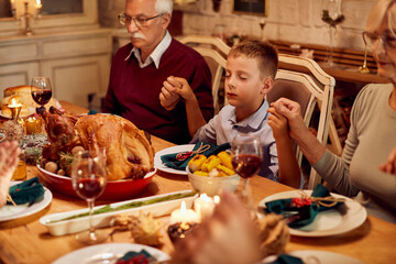 Small boy and his family blessing food while holding hands during Thanksgiving meal at dining table.