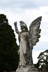Fototapeta premium Stone sculpture of an angel holding a horn in a cemetery, with bright but cloudy sky in the background
