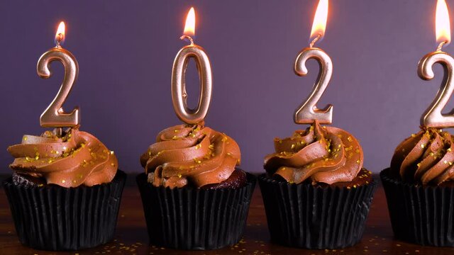 Happy New Year's Eve 2022 chocolate cupcakes decorated with gold burning candles on aqainst dark wood table setting background. Pan across.
