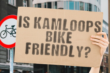 The question " Is Kamloops bike friendly? " on a banner in men's hand with blurred background. Transportation. Zero waste. Bicycle lane. Streets. City. Safety. Insecure. Road signs. Dangerous