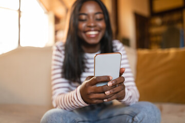 Happy black woman with smartphone browsing web, checking email, posting in social network, sending sms on couch at home
