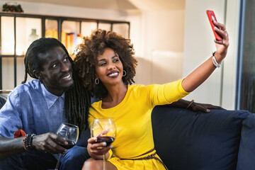 multiracial couple having fun taking smartphone's selfies at home sitting on the couch. Best friends using cellphone on the sofa. High iso image, focus on the woman.