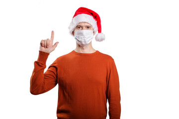 Fototapeta na wymiar an isolated shot of a happy man in a medical mask from the coronavirus, in casual clothes, an orange sweater, a Christmas hat. With his finger he points up, looks into the camera