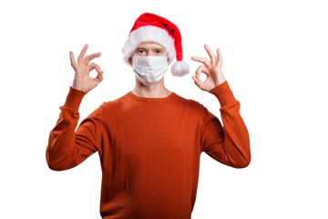 Fototapeta na wymiar an isolated shot of a happy man in a medical mask from the coronavirus, in casual clothes, an orange sweater, a Christmas hat. Shows the gesture ok, looks at the camera
