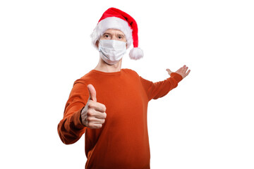 Fototapeta na wymiar an isolated shot of a happy man in a medical mask from the coronavirus, in casual clothes, an orange sweater, a Christmas hat. gesture ok, the other hand points back, looks into the camera
