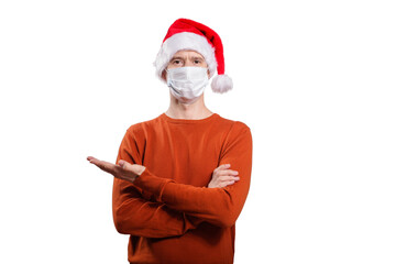 Fototapeta na wymiar an isolated shot of a happy man in a medical mask from the coronavirus, in casual clothes, an orange sweater, a Christmas hat. With his finger he points to the left, looks into the camera