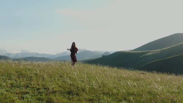 Aerial view of a young barefoot woman in a red dress who dances on the grass against the background of hills and mountains in the fog under a blue sky in the rays of the sunset. Circular panorama