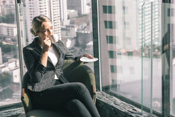 Young business woman holding a cup of coffee. She is wearing white shirt and suit. She is sitting in a hotel lobby.