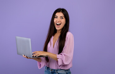 Great website. Surprised armenian woman holding laptop and exclaiming with excitement, standing over violet background