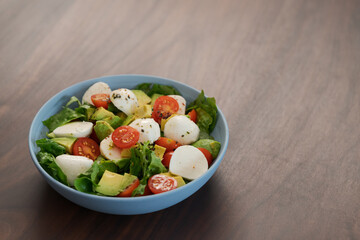 Salad with avocado, cherry tomatoes, romaine and mozzarella in blue bowl on walnut table with copy space