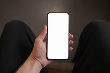 Man hold smartphone with white screen in his hand sitting on a couch in home environment