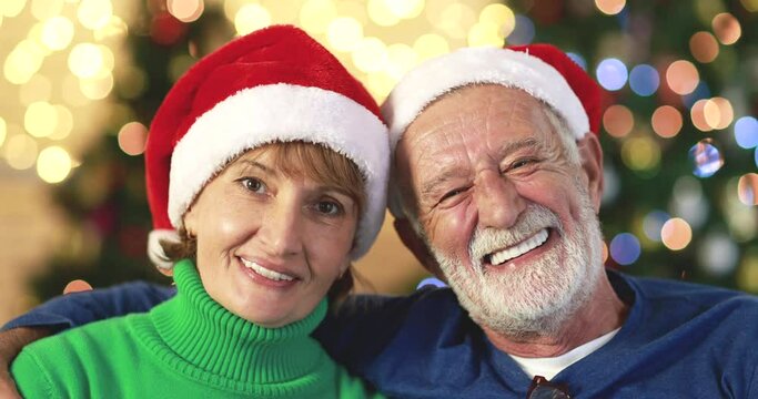 Cheerful elderly man with gray beard embracing smiling mature woman both wearing Santa hats and looking at camera sitting on sofa in room with Xmas glowing trees