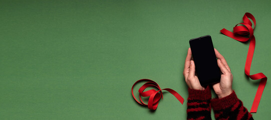 Top view.Woman wearing red sweater and holding smart phone with blank screen, red xmas ribbon on the green surface.Online buying presents,gifts