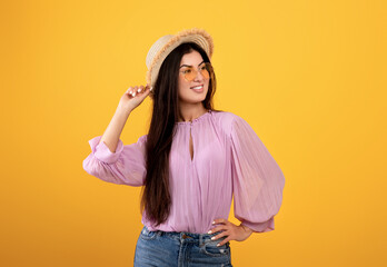 Stylish armenian lady in sunglasses, straw hat and shirt posing and smiling over yellow studio background