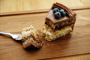 Piece of Kiev cake with chocolate icing and blueberries on wooden board. A bite of cake on a fork