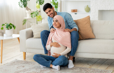 Naklejka na ściany i meble Pregnant muslim woman feeling relaxed, sitting on floor at living room and attentive husband massaging her shoulders