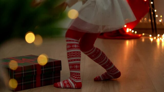 Little childrens feet in red tights with New Years ornament go to Christmas tree with gifts at dark. Child in socks puts gift under Christmas tree