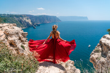 A girl with loose hair in a long red dress descends the stairs between the yellow rocks overlooking the sea. A rock can be seen in the sea. Sunny path on the sea from the rising sun