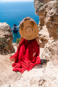 A girl with loose hair in a long red dress descends the stairs between the yellow rocks overlooking the sea. A rock can be seen in the sea. Sunny path on the sea from the rising sun