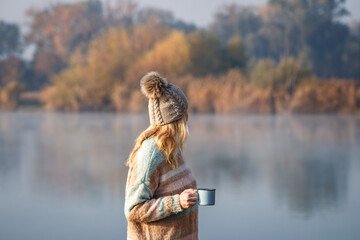 Woman relaxing with coffee next to lake at autumn cold morning. Camping and hiking outdoors at fall...