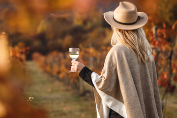 Female vintner enjoying white wine in her vineyard. Woman with poncho and hat holding glass of wine...