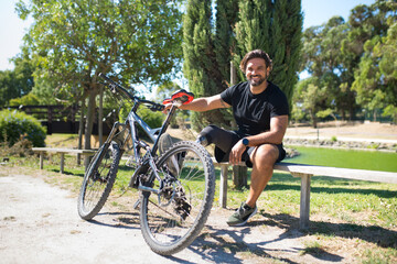 Fototapeta premium Smiling man with disability with bicycle. Caucasian man with mechanical leg sitting on bench, resting after training, looking at camera. Sport, leisure concept