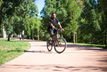 Obraz premium Person with disability riding bicycle. Curly man with mechanical leg exercising in park on sunny day. Sport, disability concept