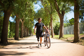 Front view of person with disability with bicycle. Curly man with mechanical leg walking in park on sunny day. Sport, disability concept