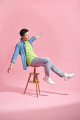 Asian man in stylish clothes sitting on high stool on pink background