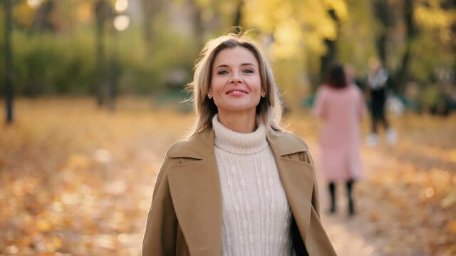 Attractive mature blond woman walking down the park turns around with flying hair and looking at the camera outdoors Happy relaxed female walking on the autumn park enjoying beautiful time