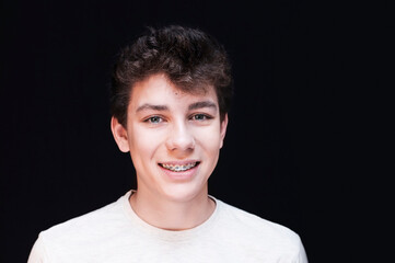 Cute teenager guy on a dark background in a white T-shirt smiling, clearly visible braces on his teeth to align teeth. Dental concept
