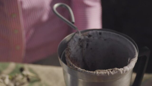 woman in a pink striped shirt puts two tablespoons of coffee in a rustic coffee pot with a cloth strainer