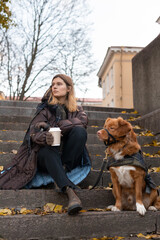 Beauty Caucasian girl in brown coat and with cup of coffee sitting with Nova Scotia Duck Tolling Retriever on stone steps. Outdoor leisure with dog. Selective focus. Pets loving concept.