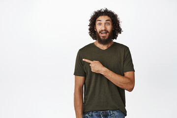 Happy young man looking surprised, pointing finger left, showing advertisement, standing in t-shirt...