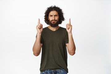 Skeptical middle easter man pointing fingers up, frowning and looking disappointed, express dislike, standing in t-shirt over white background