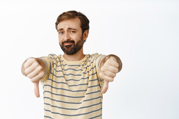 Fototapeta premium Very bad. Portrait of bearded guy shows thumbs down and smirks with dislike, disappointed by smth, standing over white background