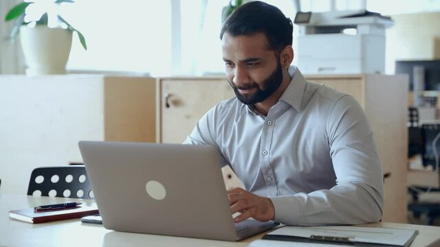 Ethnic indian business man entrepreneur investor manager using computer, watching webinar working in office analyzing online data market thinking doing web research looking at laptop.