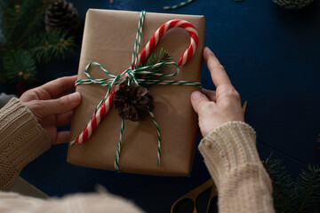 woman wraps a handmade eco style christmas present in kraft paper and natural spruce branches on a blue wooden background.