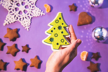 christmas decoration in hand. green toy tree in hands. christmas tree decorations, gingerbread and fruits on purple table