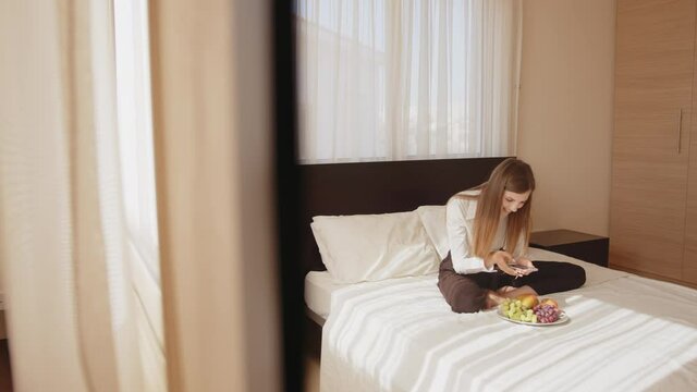 Happy young woman sitting on the bed with a plate of fresh fruits and taking pictures on a mobile phone. Woman traveler enjoying free time in a cozy hotel room. People and technology concept.