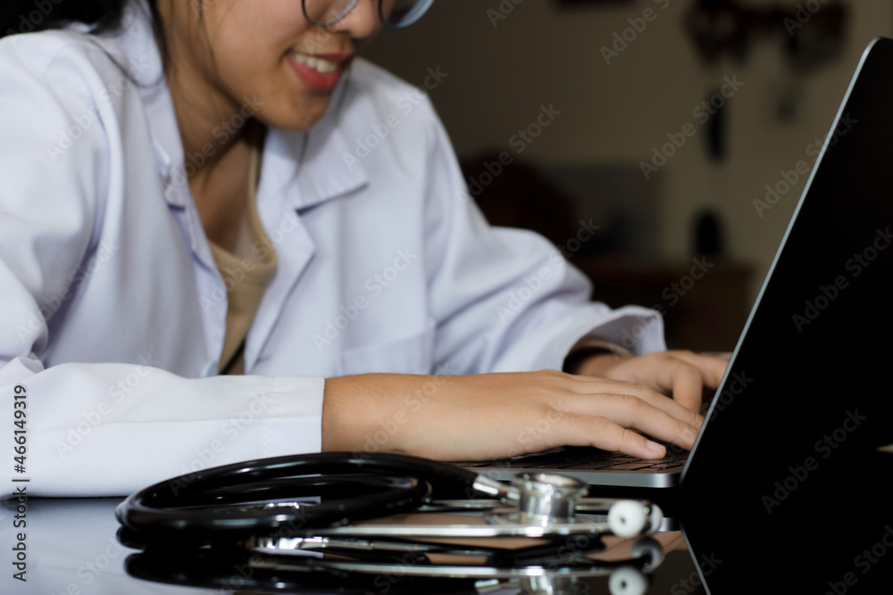 Wall mural Young smiling female doctor or medical student with eyeglasses working on laptop computer with stethoscope on the desk at medical office. 