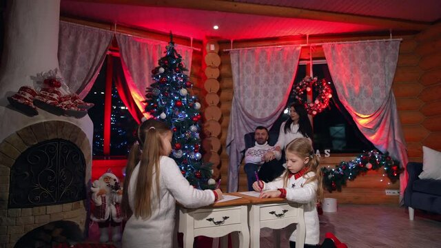 Cozy atmosphere at Christmas in the house. Happy parents talk together while little girls painting in the Christmas decorated room.