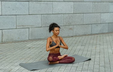 Tuinposter Positive lady in stylish sportswear meditates sitting in lotus pose on mat against grey stone wall at training © sergiymolchenko