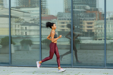 Fototapeta premium Pretty sportive lady in tracksuit holds hands on waist standing against modern building with metal fence gate in sunny city