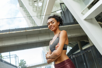 Concentrated lady in stylish top with crossed arms stands against contemporary city building fence