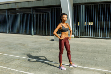 Pretty sportive lady in tracksuit holds hands on waist standing against modern building with metal fence gate in sunny city